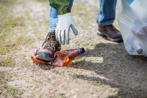 A person wearing white gloves and blue jeans is seen picking up a used, partially crushed amber glass bottle from a gravel or dirt surface outside. The bottle is lying on the ground and appears to be an empty beverage container with a dark cap. Nearby, a transparent plastic bag filled with waste is visible on the right side of the image. The background includes a grassy area, suggesting an outdoor setting consistent with a property or garden where rubbish might be collected or prepared for disposal. The scene emphasizes waste collection and disposal activity relevant to commercial waste removal services offered by companies like Commercial Waste Removal Mitcham, serving local areas around Mitcham and the postcode area. The lighting is natural, with clear daylight illuminating the scene, focusing on proper handling and collection of recyclable or rubbish materials in compliance with local waste management practices.