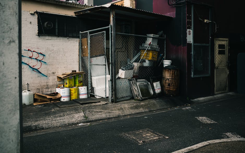 A collection of black garbage bags, cardboard boxes, and loose waste materials piled up in front of a red metal fire door set into a rough-textured, off-white concrete wall. The door is covered with g